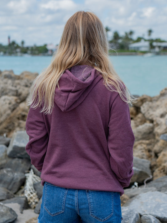 Woman wearing a mauve hoodie, standing by a waterfront with rocks in the background.