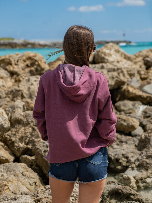 Woman wearing a mauve hoodie, standing by a waterfront with rocks in the background.