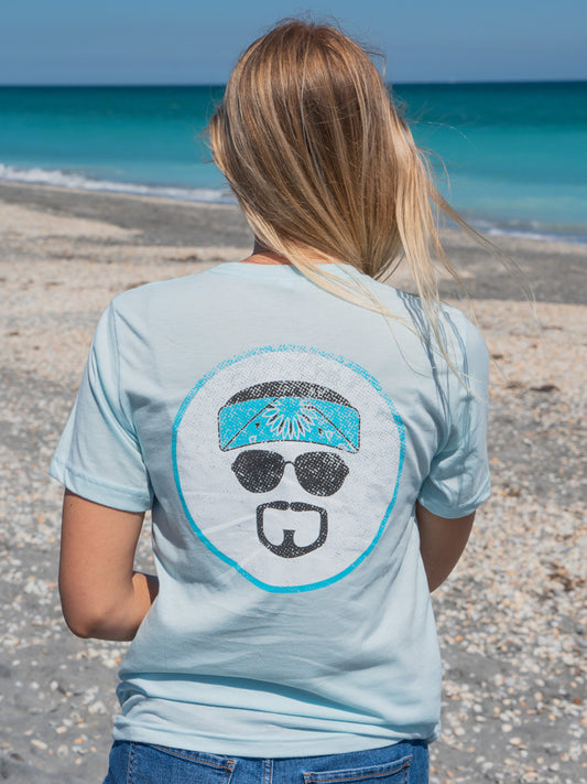 Woman standing on a beach wearing a light blue t-shirt with a logo, looking at the ocean.