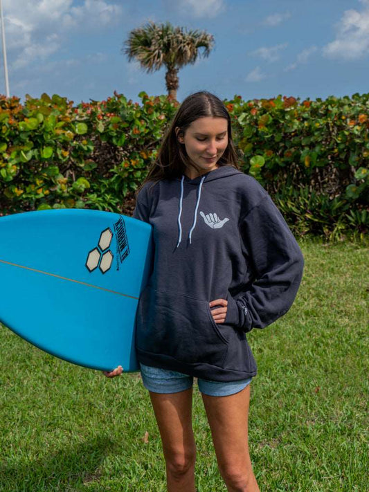 Woman holding a surfboard wearing dark grey hoodie with a hang loose sign on the front with greenery in the background