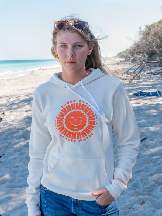 Woman wearing white hoodie with sun design, standing on a beach with trees in the background