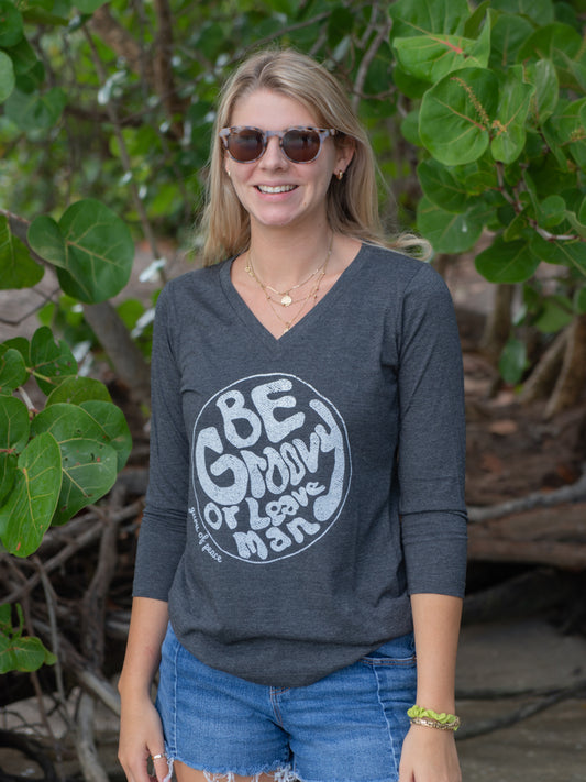 Woman wearing a gray long-sleeve shirt with a graphic design by a beach with leafy background.