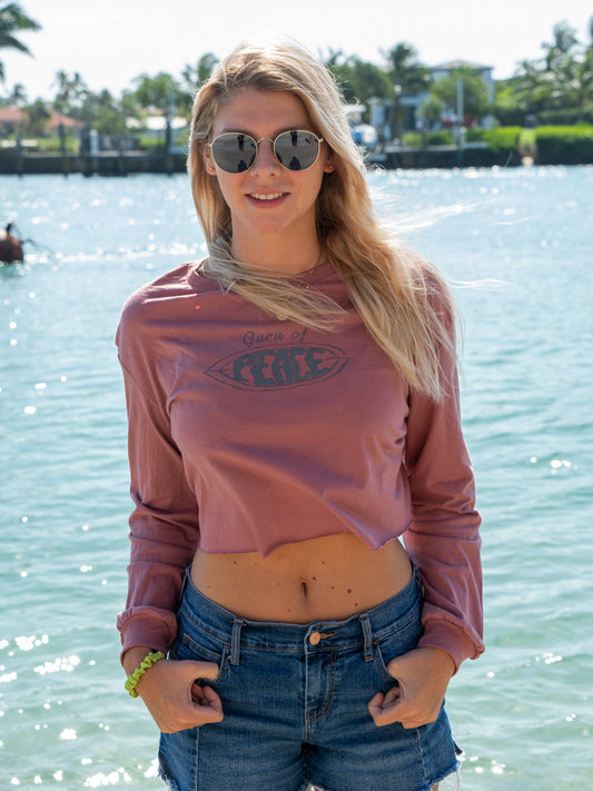 Woman wearing a mauve long-sleeve crop top with a logo and text, standing by a body of water