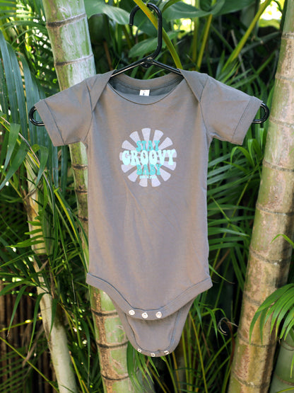 Asphalt colored baby onesie with 'Stay Groovy Baby' text on a hook hanging on a tree