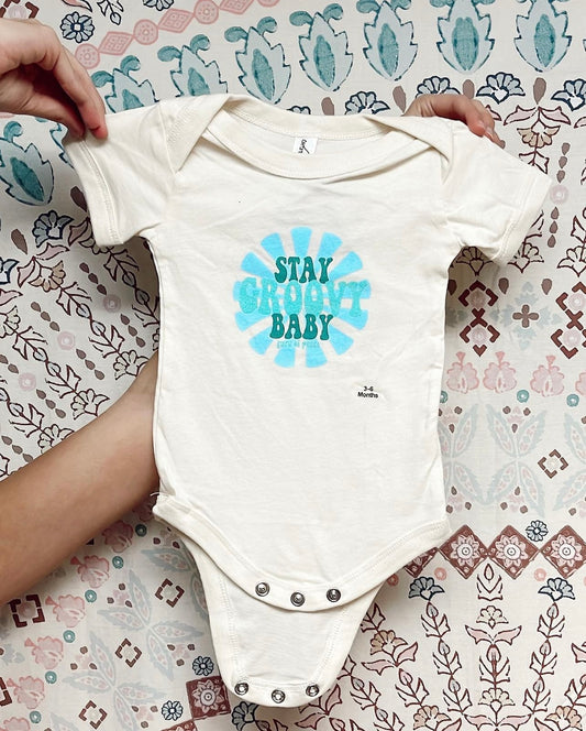 White baby onesie with 'Stay Groovy Baby' text held by a hand on a patterned background