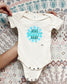 White baby onesie with 'Stay Groovy Baby' text held by a hand on a patterned background
