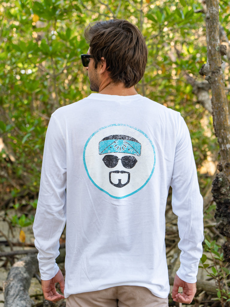 Man wearing a white long-sleeve shirt with logo, standing outdoors among greenery.