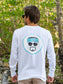 Man wearing a white long-sleeve shirt with logo, standing outdoors among greenery.