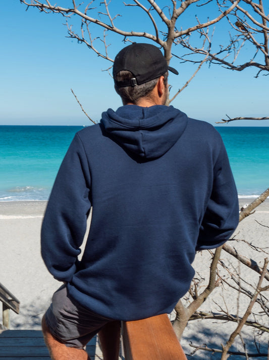 Man wearing navy hoodie, sitting on a wooden bench in a beach setting