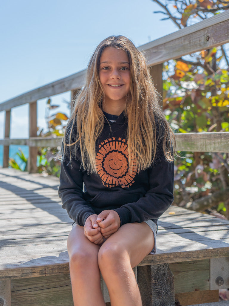 Young girl wearing black hoodie with a sun design, sitting on a wooden bench outdoors.