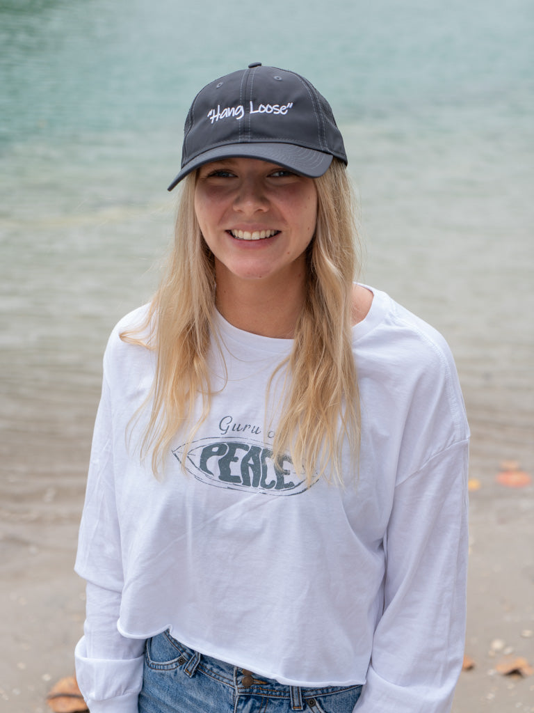Person wearing a gray sports cap with the words 'Hang Loose' and white long-sleeve shirt with 'Peace' graphic on a beach