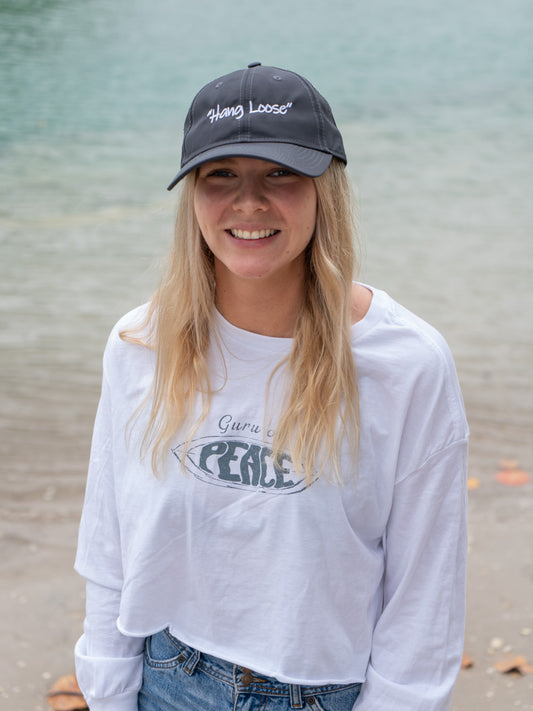 Person wearing a gray sports cap with the words 'Hang Loose' and white long-sleeve shirt with 'Peace' graphic on a beach