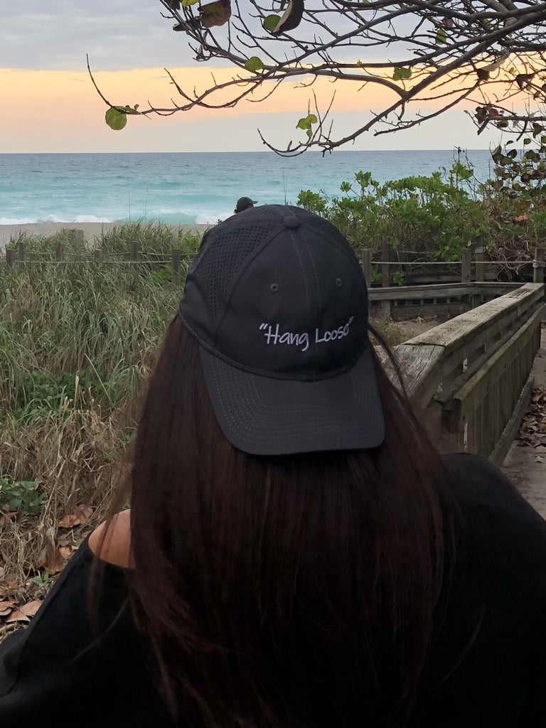 A person sitting on a beach with a backwards black hat that has the words 'Hang Loose' on it
