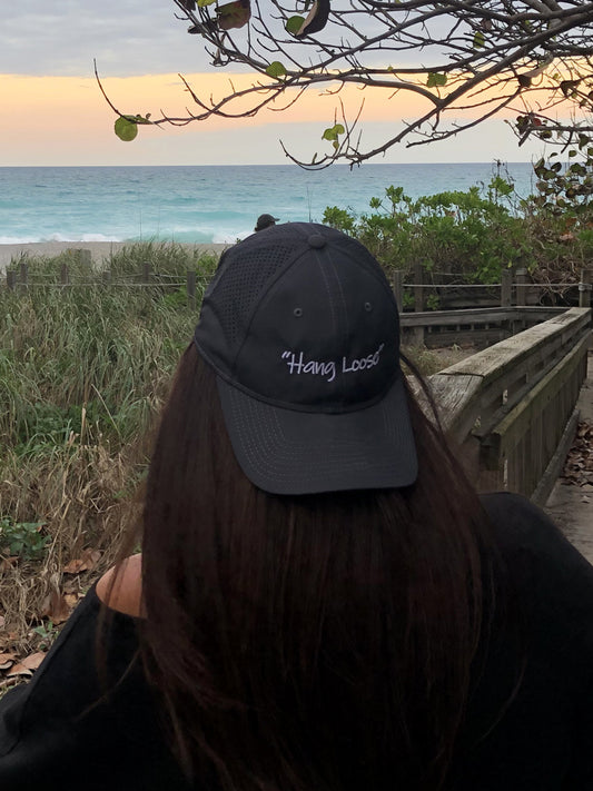 A person sitting on a beach with a backwards black hat that has the words 'Hang Loose' on it
