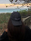 A person sitting on a beach with a backwards black hat that has the words 'Hang Loose' on it