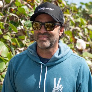 Person wearing a blue hoodie and black cap with sunglasses, standing against a green leafy background.