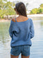 Woman wearing a wide neck blue crewneck by a body of water