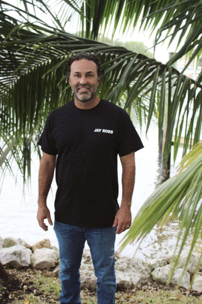 Man wearing a black t-shirt with 'JAY ROBB' text, standing in front of palm trees.