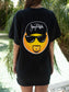 Woman wearing black t-shirt with an emoji on the back, standing by a body of water.