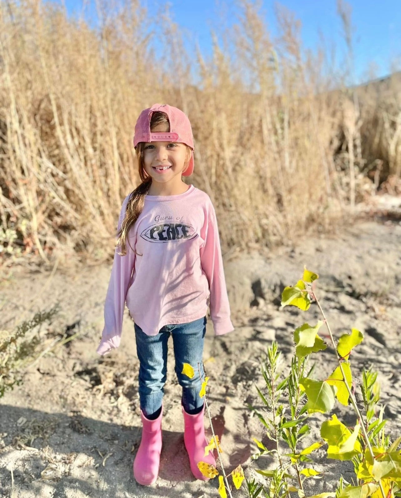 Young girl in a pink 'Guru of Peace' shirt holding a flower, standing on rocks with a blue sky background.