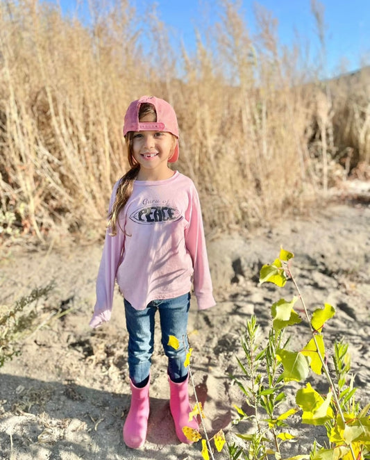 Young girl in a pink 'Guru of Peace' shirt holding a flower, standing on rocks with a blue sky background.