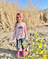 Young girl in a pink 'Guru of Peace' shirt holding a flower, standing on rocks with a blue sky background.