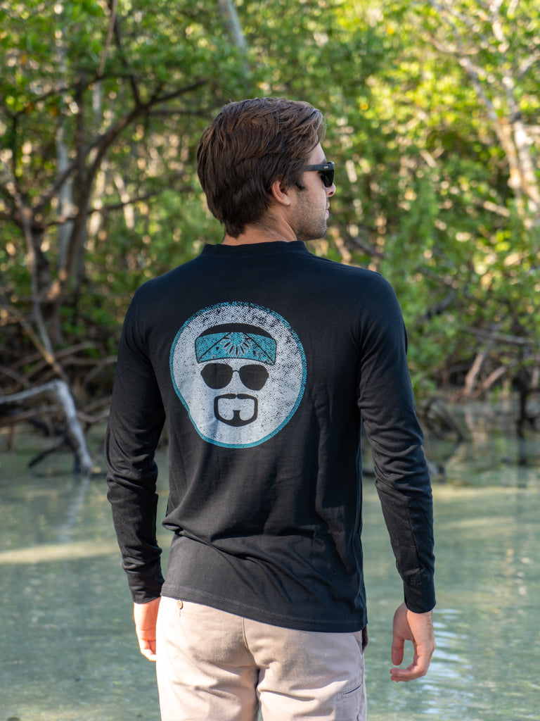 Man wearing a black long-sleeve shirt with logo, standing outdoors on a beach by water with trees in the background.