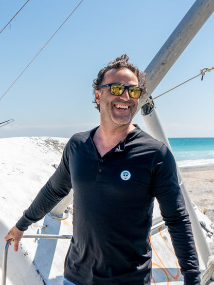 Man wearing a black long-sleeve shirt with logo, standing outdoors on a beach on a boat.
