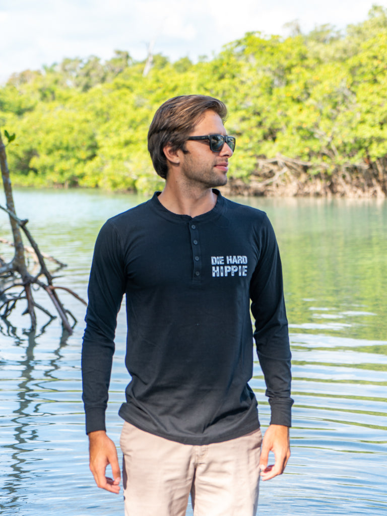 Man wearing a black long-sleeve shirt with text, standing outdoors on a beach by  water.