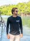 Man wearing a black long-sleeve shirt with text, standing outdoors on a beach by  water.