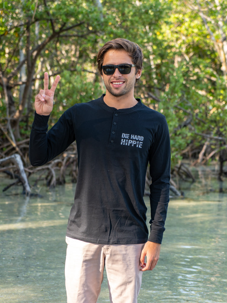 Man wearing a black long-sleeve shirt with text, standing outdoors on a beach by water with trees in the background.