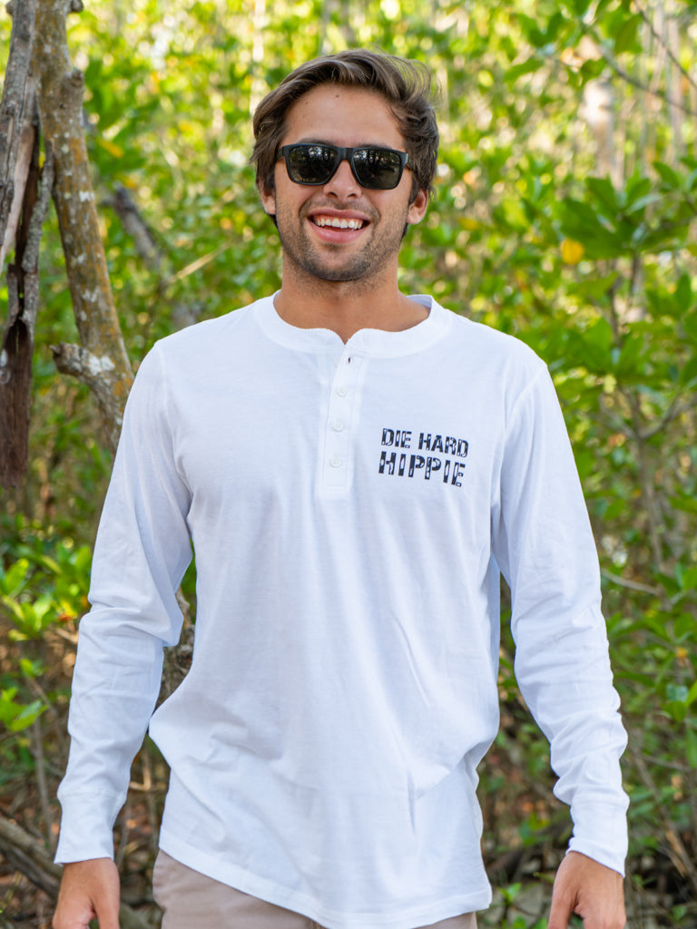 Man wearing a white long-sleeve shirt with text, standing outdoors among greenery.