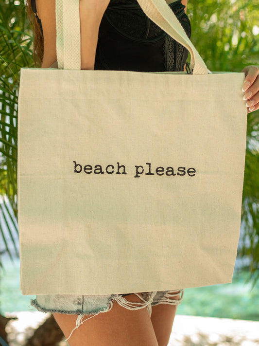 Person holding a beige tote bag with 'beach please' text against a blurred natural background