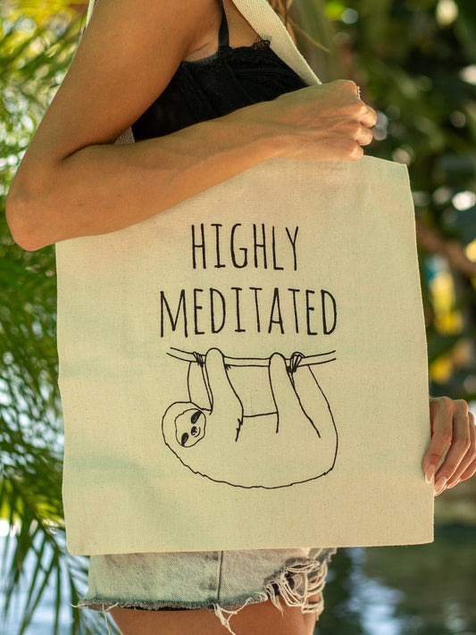 Person holding a beige tote bag with 'highly meditated' text against a blurred natural background