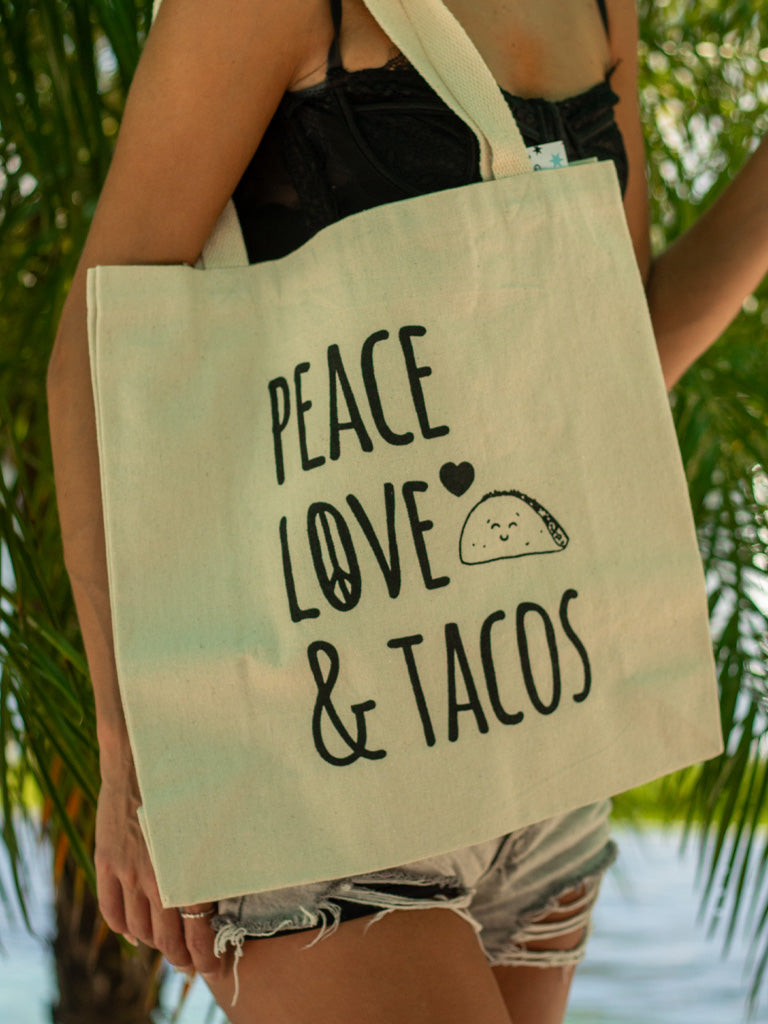 Person holding a beige tote bag with 'peace love & tacos' text against a blurred natural background