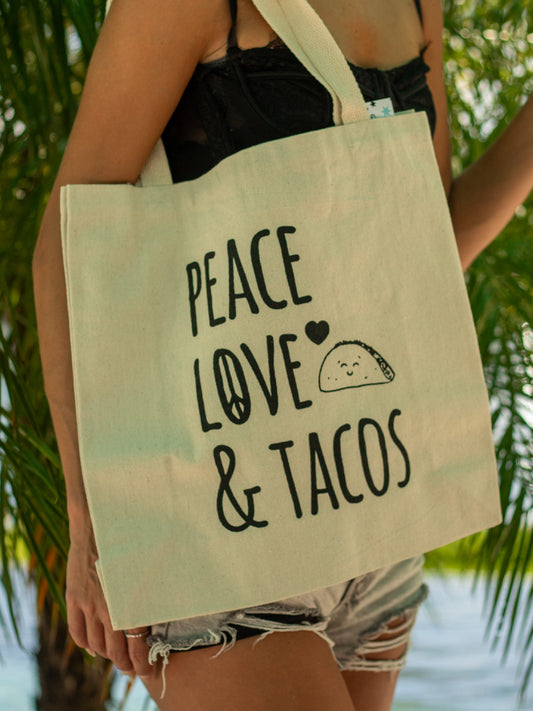 Person holding a beige tote bag with 'peace love & tacos' text against a blurred natural background