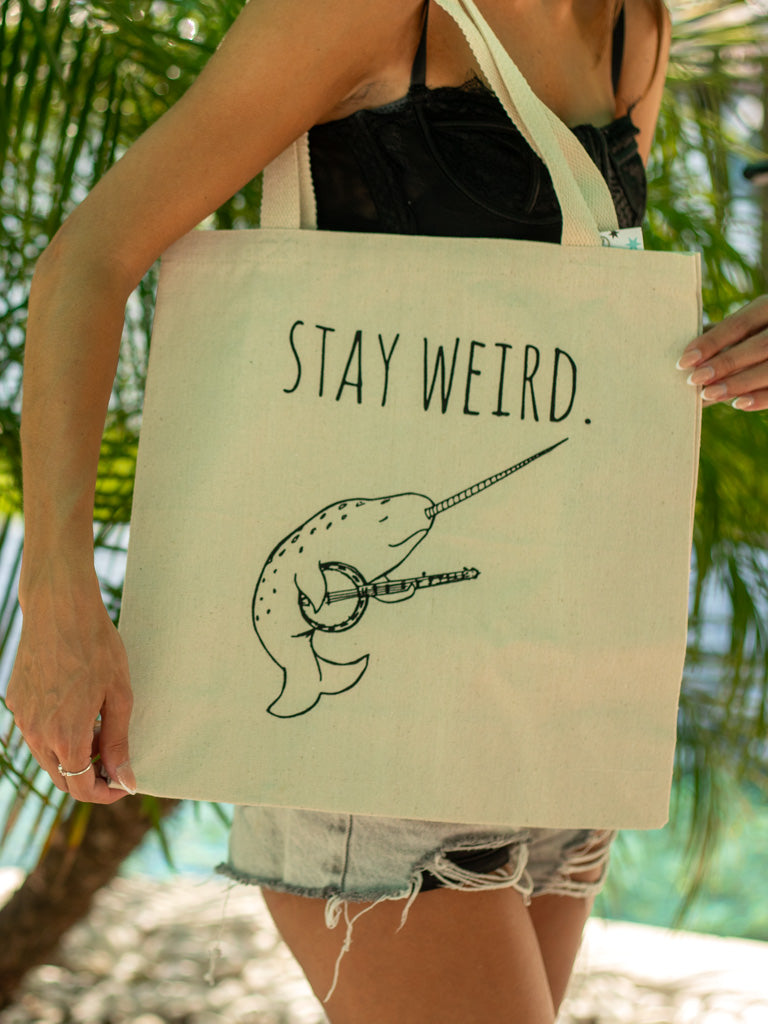 Person holding a beige tote bag with 'stay weird' text against a blurred natural background