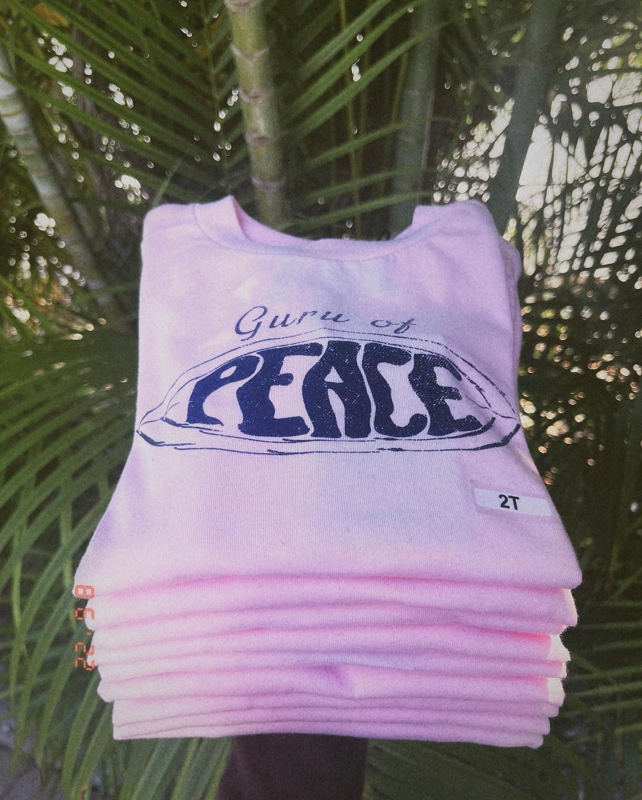 Pink 'Guru of Peace' shirts stacked with palm tree leaves in the background.