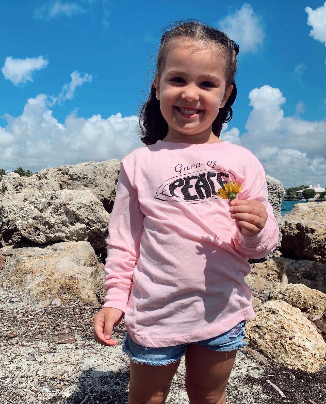 Young girl in a pink 'Guru of Peace' shirt holding a flower, standing on rocks with a blue sky background.