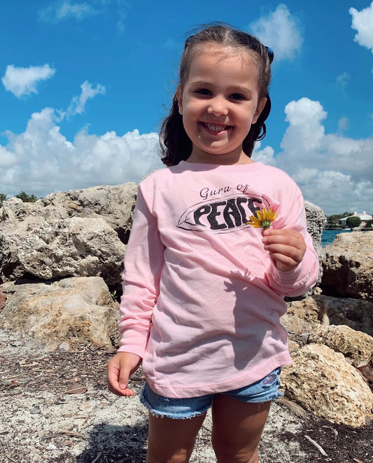 Young girl in a pink 'Guru of Peace' shirt holding a flower, standing on rocks with a blue sky background.