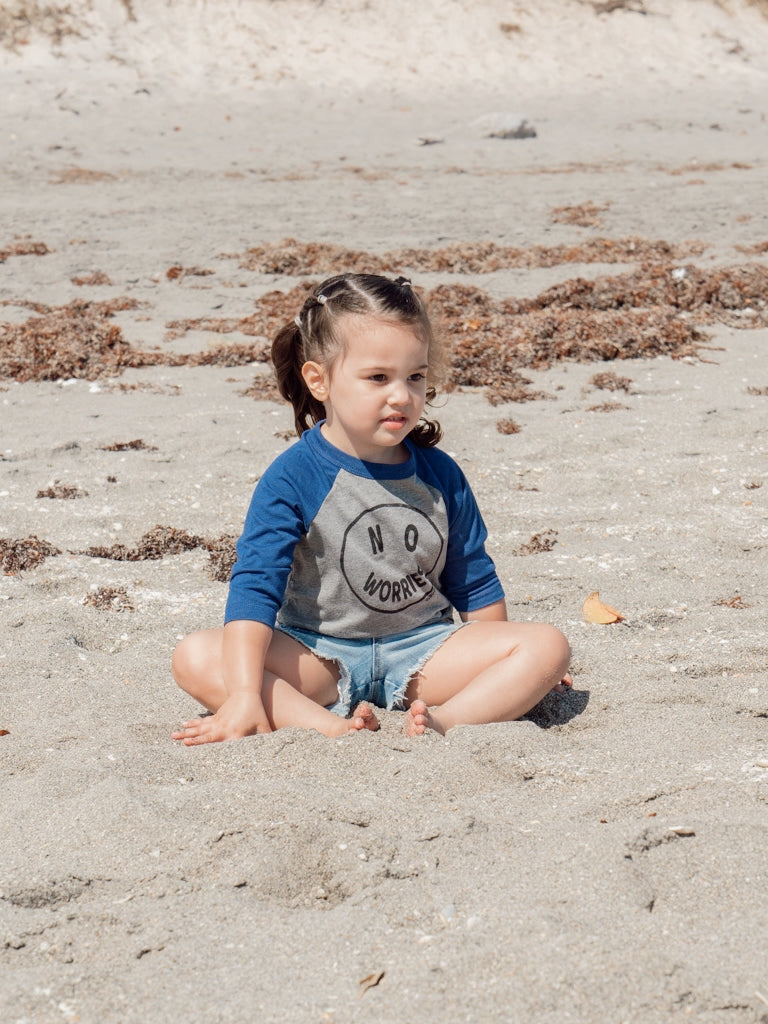 Child sitting on the beach with a basbeall tee that say 'No Worries'