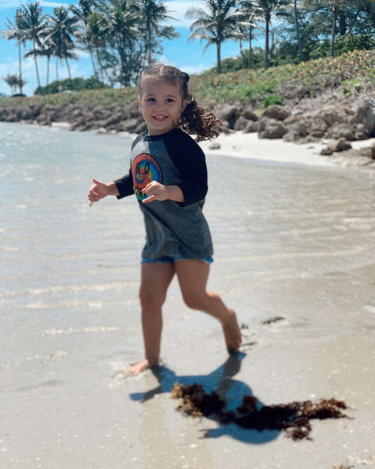 Child in a grey shirt with a colorful design standing on a sandy beach with palm trees in the background