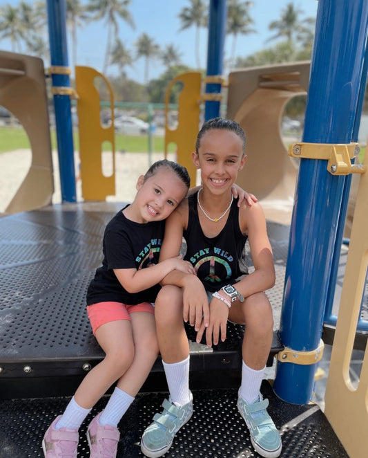 Two young girls sitting on a playground bench wearing matching 'Stay Wild' shirts.