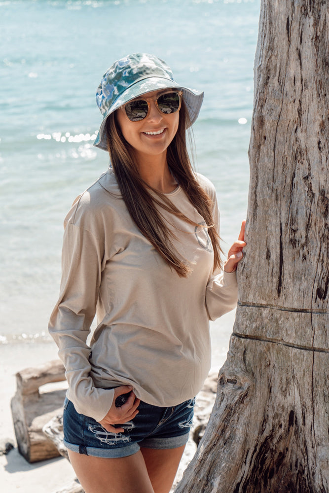 Woman standing next to a tree wearing a tan long-sleeve shirt with a peace symbol and denim shorts.