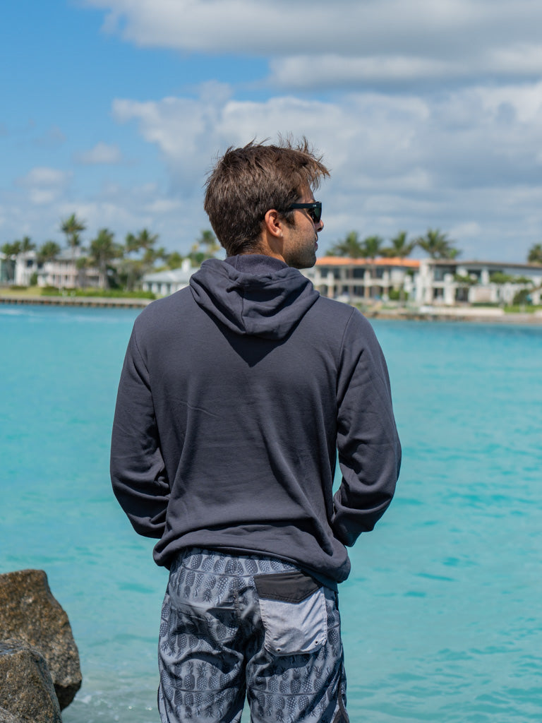 Man wearing a dark gray hoodie, standing by a waterfront with buildings and trees in the background.