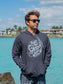 Man wearing a dark gray hoodie with a logo and text, standing by a waterfront with buildings and trees in the background.