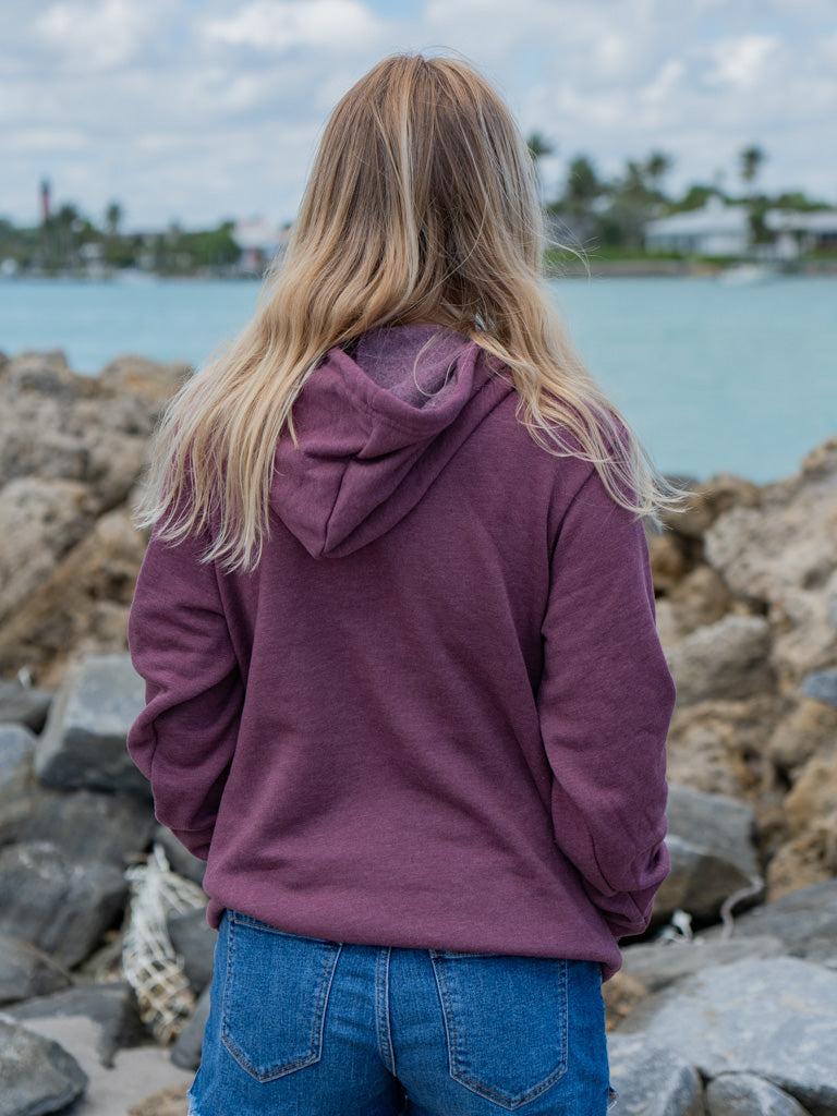 Woman wearing a mauve hoodie, standing by a waterfront with rocks in the background.