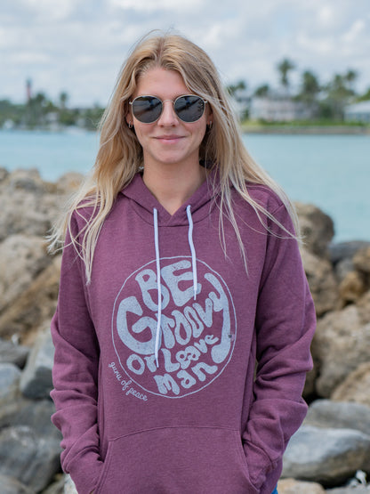 Woman wearing a mauve hoodie with a logo and text, standing by a waterfront with rocks and buildings in the background.