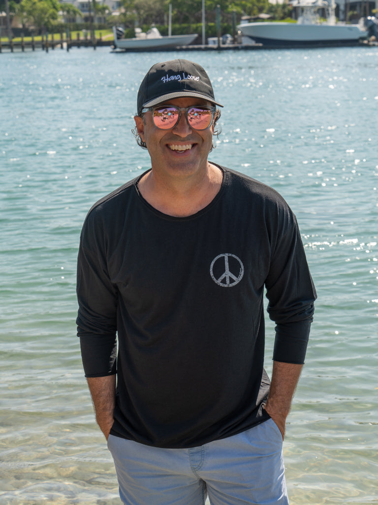 Man standing by a body of water wearing a black long-sleeve shirt with a peace symbol.