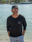 Man standing by a body of water wearing a black long-sleeve shirt with a peace symbol.